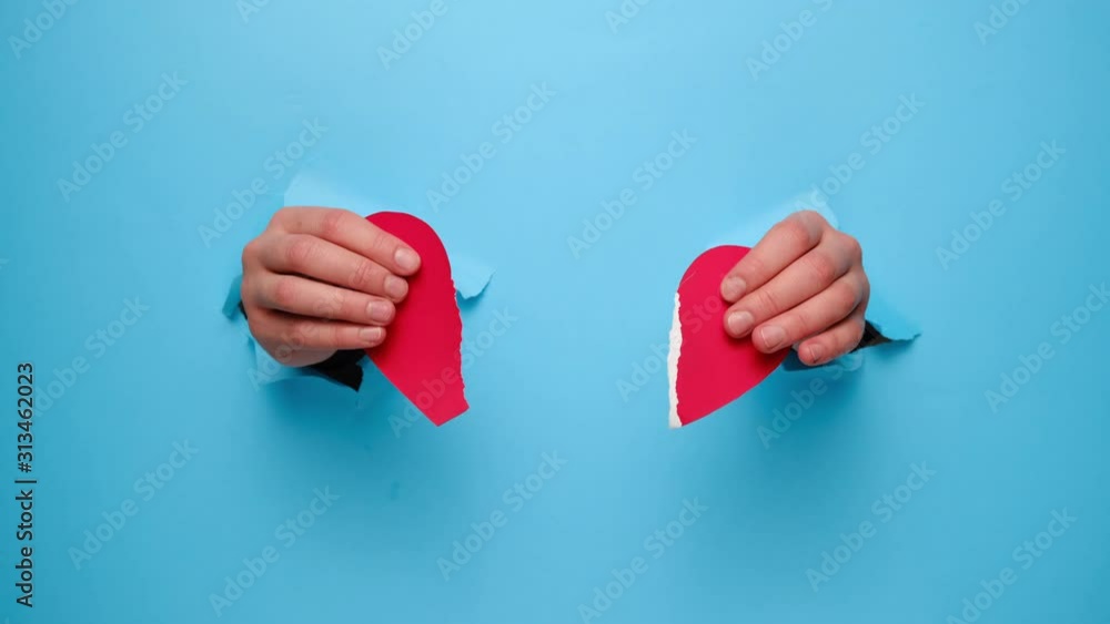 Female hands breaks a red paper heart through a torn blue paper wall. Heart broken, Love and Valentines day concept.