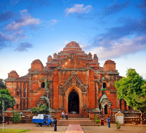 People in front of a temple surrounded by green vegetation in old Bagan, Myanmar, under a colorful sunrise sky.
