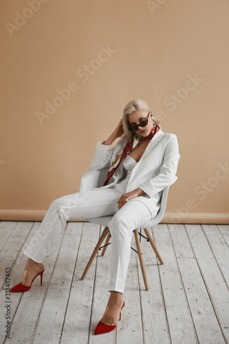 Beautiful young woman with perfect blond hair in elegant white suit and red shoes posing in studio. Fashionable blonde model girl in stylish outfit and sunglasses. Lady boss ready for office meeting