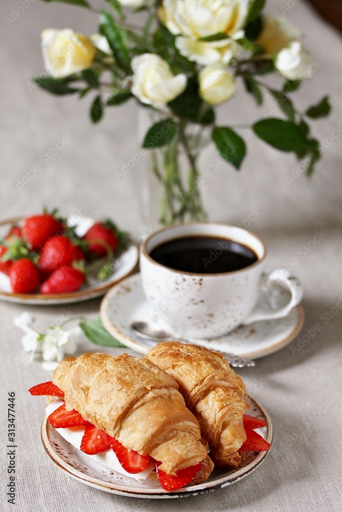 Breakfast in French. Croissant sandwich with I soft cheese and strawberries. And black coffee, a bouquet of flowers. Fresh strawberries. Soft focus. Space for text. Copy Space Breakfast in French. Croissant sandwich with I soft cheese and strawberries. And black coffee, a bouquet of flowers. Fresh strawberries. Soft focus. Space for text. Copy Space