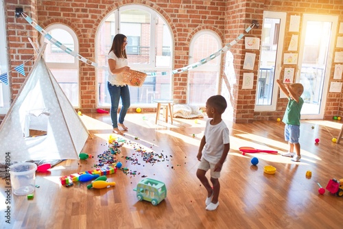 Young beautiful teacher and toddlers playing basketball around lots of toys at kindergarten