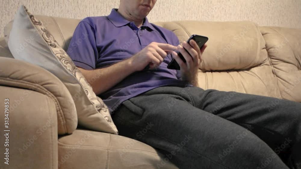 A man is lying on the couch and uses a smartphone-is in social networks, flipping through photos or running a business from the comfort of home, ordering goods or paying for online purchases. A man is lying on the couch and uses a smartphone-is in social networks, flipping through photos or running a business from the comfort of home, ordering goods or paying for online purchases.