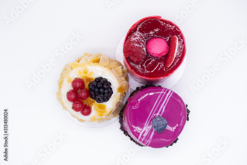 Three cakes on a white background: Bright red with raspberries, macaroon on top, cream cake with almond petals, red currants and blackberries and pink mousse with blueberries and chocolate chips