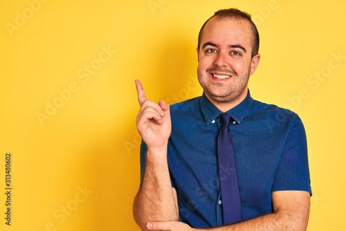 Young businessman wearing denim casual shirt and tie over isolated yellow background with a big smile on face, pointing with hand and finger to the side looking at the camera.