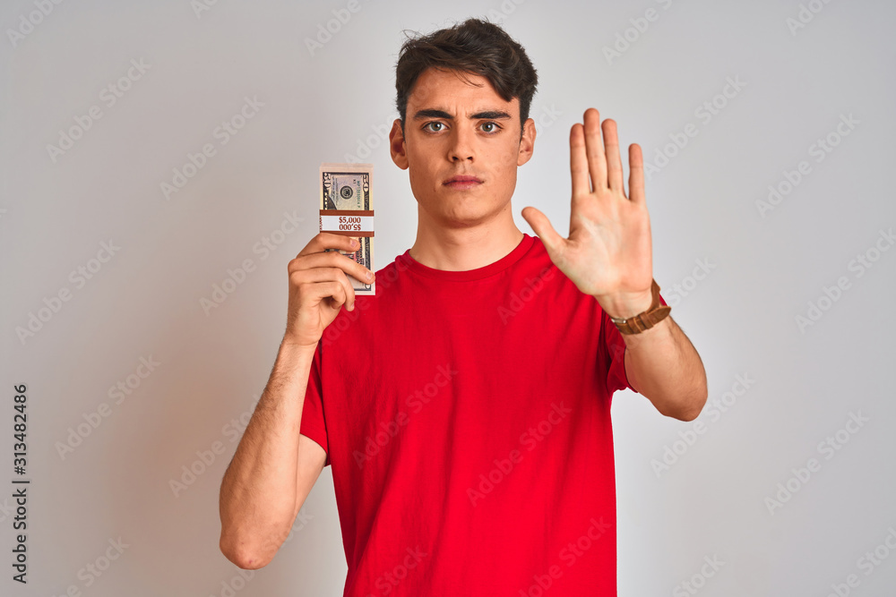 Teenager boy holding a bunch of dollars bank notes over isolated background with open hand doing stop sign with serious and confident expression, defense gesture Teenager boy holding a bunch of dollars bank notes over isolated background with open hand doing stop sign with serious and confident expression, defense gesture