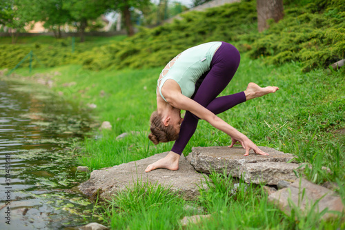 A young sports girl practices yoga on a green lawn by the river, yoga assans posture. Meditation and unity with nature