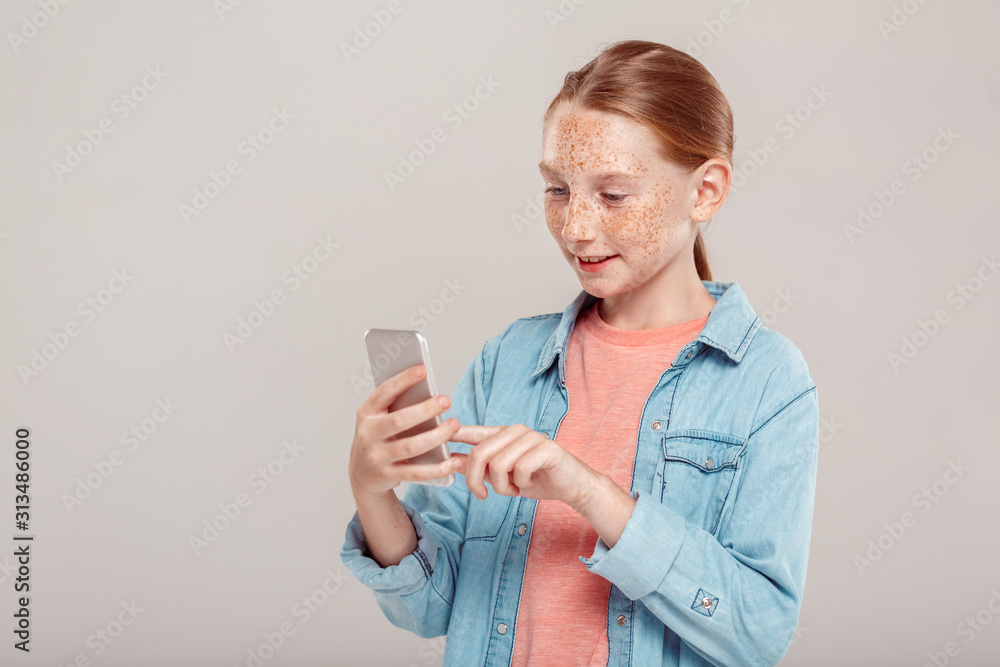 Inclusive Beauty. Girl with freckles in denim jacket standing isolated on grey browsing smartphone smiling curious