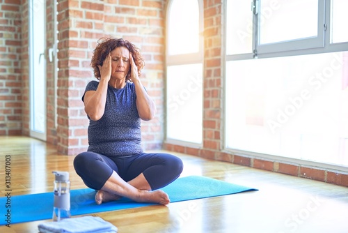 Middle age beautiful sportswoman wearing sportswear sitting on mat practicing yoga at home with hand on head for pain in head because stress. Suffering migraine.