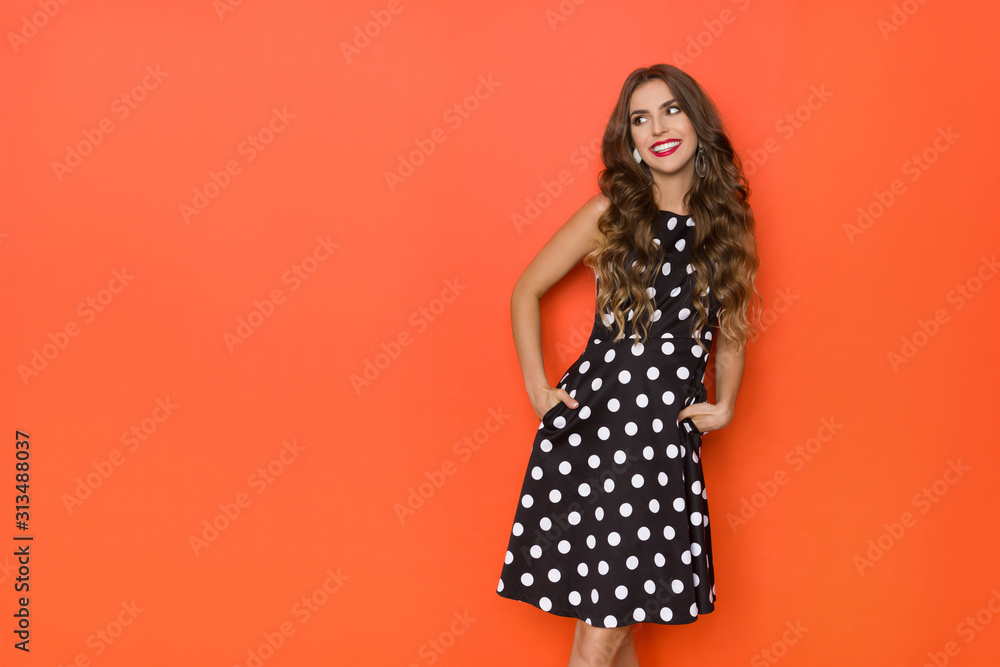 Cute Young Woman In Black Coctail Dress In Polka Dots Is Holding Hands In Pockets, Looking Away And Smiling. Cute Young Woman In Black Coctail Dress In Polka Dots Is Holding Hands In Pockets, Looking Away And Smiling.