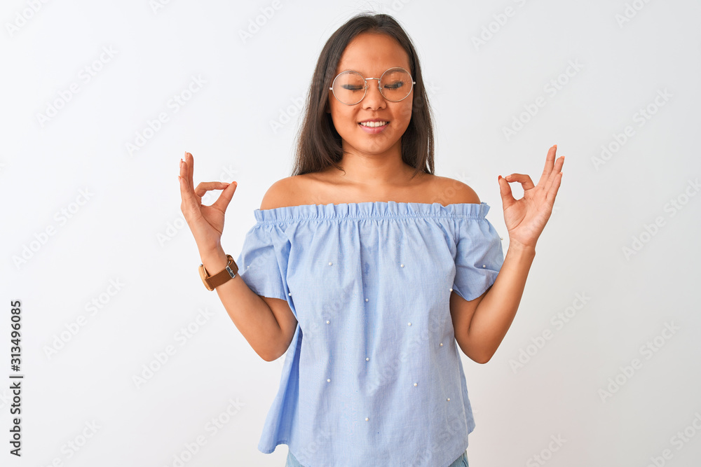 Young chinese woman wearing blue t-shirt and glasses over isolated white background relax and smiling with eyes closed doing meditation gesture with fingers. Yoga concept. Young chinese woman wearing blue t-shirt and glasses over isolated white background relax and smiling with eyes closed doing meditation gesture with fingers. Yoga concept.