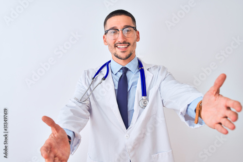 Young doctor man wearing stethoscope over isolated background looking at the camera smiling with open arms for hug. Cheerful expression embracing happiness.