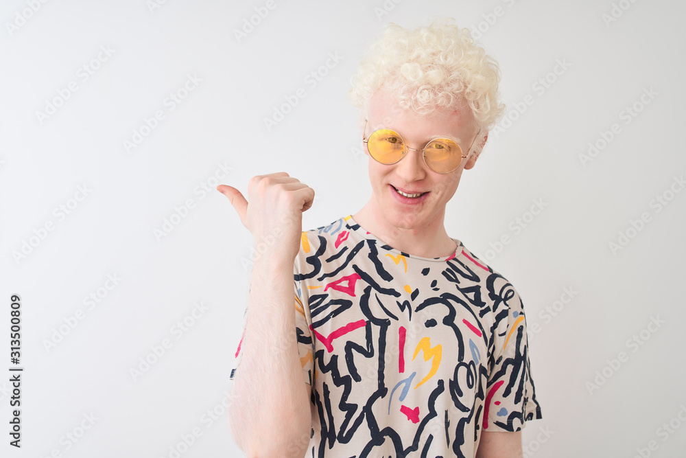 Young albino blond man wearing colorful t-shirt and sunglasses over isolated red background smiling with happy face looking and pointing to the side with thumb up. Young albino blond man wearing colorful t-shirt and sunglasses over isolated red background smiling with happy face looking and pointing to the side with thumb up.