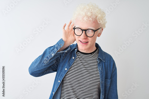 Young albino blond man wearing denim shirt and glasses over isolated white background smiling with hand over ear listening an hearing to rumor or gossip. Deafness concept.