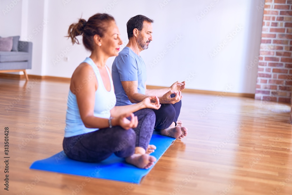 Obraz premium Middle age beautiful sporty couple sitting on mat practicing yoga doing lotus pose at gym