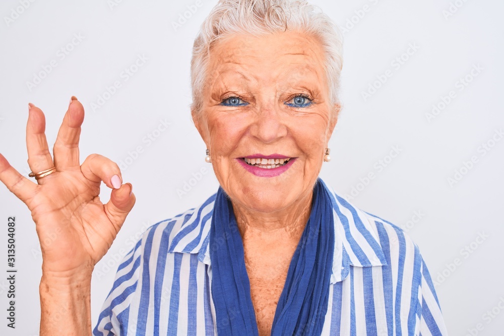 Senior grey-haired woman wearing blue striped shirt standing over isolated white background doing ok sign with fingers, excellent symbol