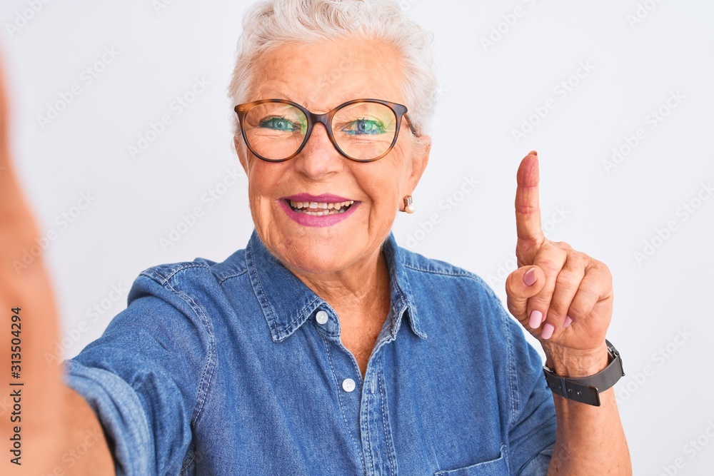 Grey-haired woman wearing denim shirt and glasses make selfie over isolated white background surprised with an idea or question pointing finger with happy face, number one Grey-haired woman wearing denim shirt and glasses make selfie over isolated white background surprised with an idea or question pointing finger with happy face, number one