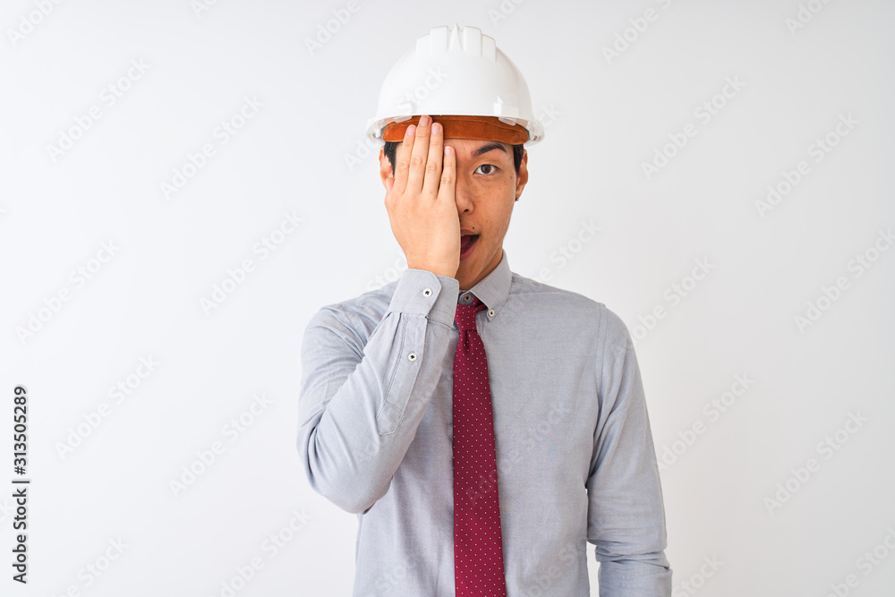 Chinese architect man wearing tie and helmet standing over isolated white background covering one eye with hand, confident smile on face and surprise emotion. Chinese architect man wearing tie and helmet standing over isolated white background covering one eye with hand, confident smile on face and surprise emotion.