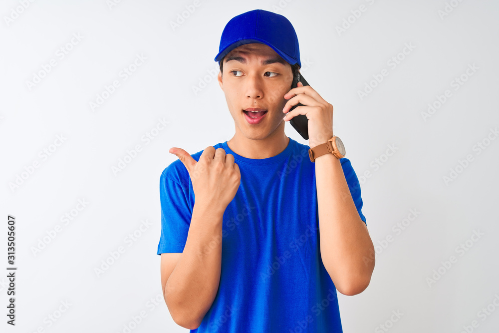 Chinese deliveryman wearing cap talking on the smartphone over isolated white background pointing and showing with thumb up to the side with happy face smiling