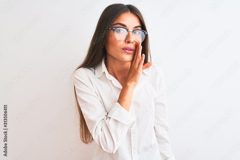 Young beautiful businesswoman wearing glasses standing over isolated white background hand on mouth telling secret rumor, whispering malicious talk conversation