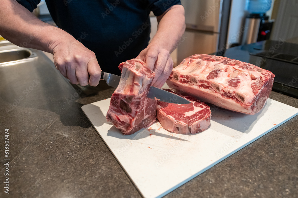 The chef uses a long stainless steel knife to cut steaks from a large prime rib roast.  The beef is on a large white plastic cutting board. Roast, steak and large pieces of meat being cut into steaks.