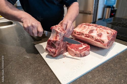 The chef uses a long stainless steel knife to cut steaks from a large prime rib roast. The beef is on a large white plastic cutting board. Roast, steak and large pieces of meat being cut into steaks.
