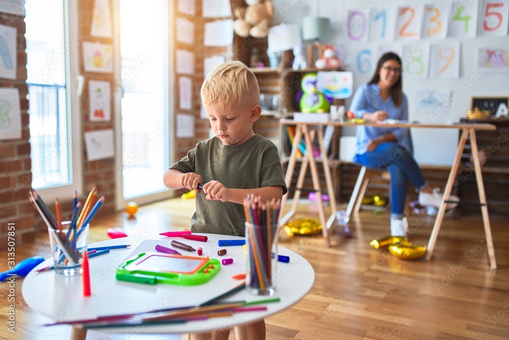 Young caucasian child playing at playschool with teacher. Mother and son at playroom drawing a draw with color pencils, young woman at the background sitting on desk. Young caucasian child playing at playschool with teacher. Mother and son at playroom drawing a draw with color pencils, young woman at the background sitting on desk.
