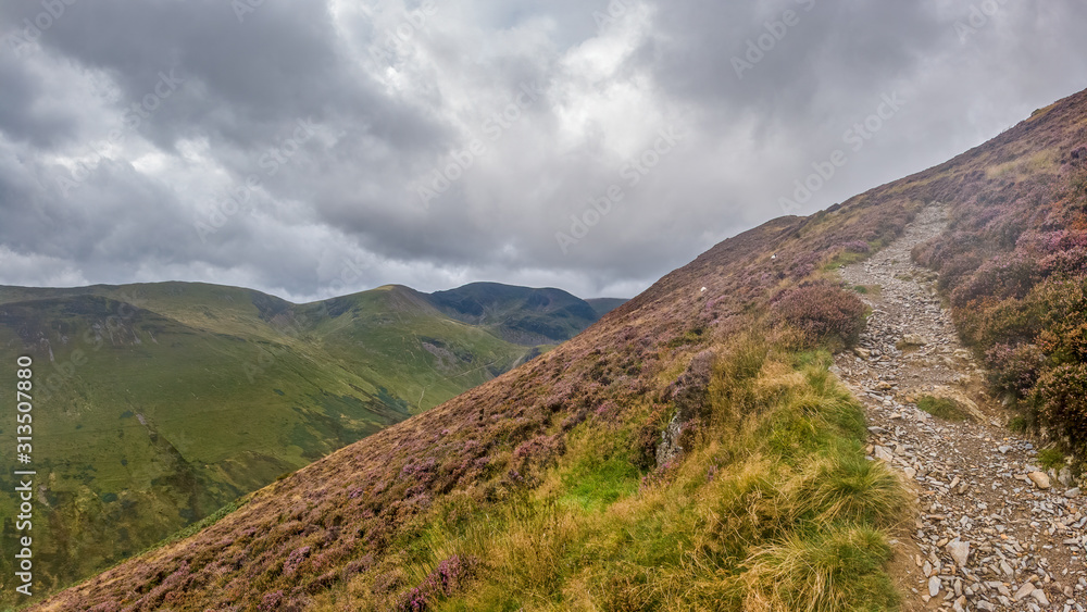 A scenic view of a mountain trail track along a grassy mountain slope and valley with mountain range in the background under a stormy grey cloudy sky A scenic view of a mountain trail track along a grassy mountain slope and valley with mountain range in the background under a stormy grey cloudy sky