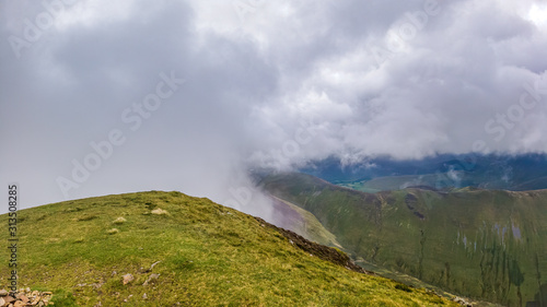 A scenic view of a grassy mpountain summit with grassy slopes and mountain range in the background under a stormy grey cloudy sky