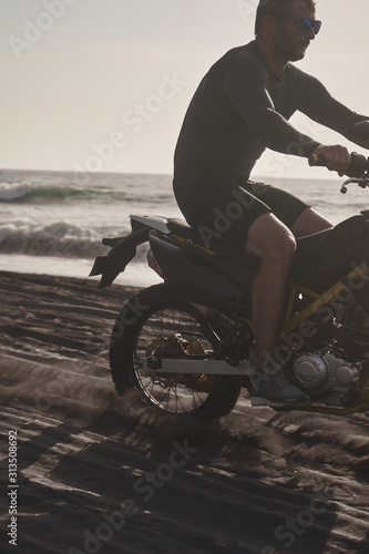 Fototapeta Naklejka Na Ścianę i Meble -  Male masculine model with cross road motorbike at the ocean coast with black sand at the golden hour time
