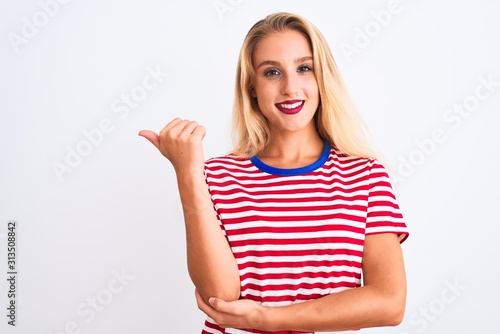 Young beautiful woman wearing red striped t-shirt standing over isolated white background smiling with happy face looking and pointing to the side with thumb up.