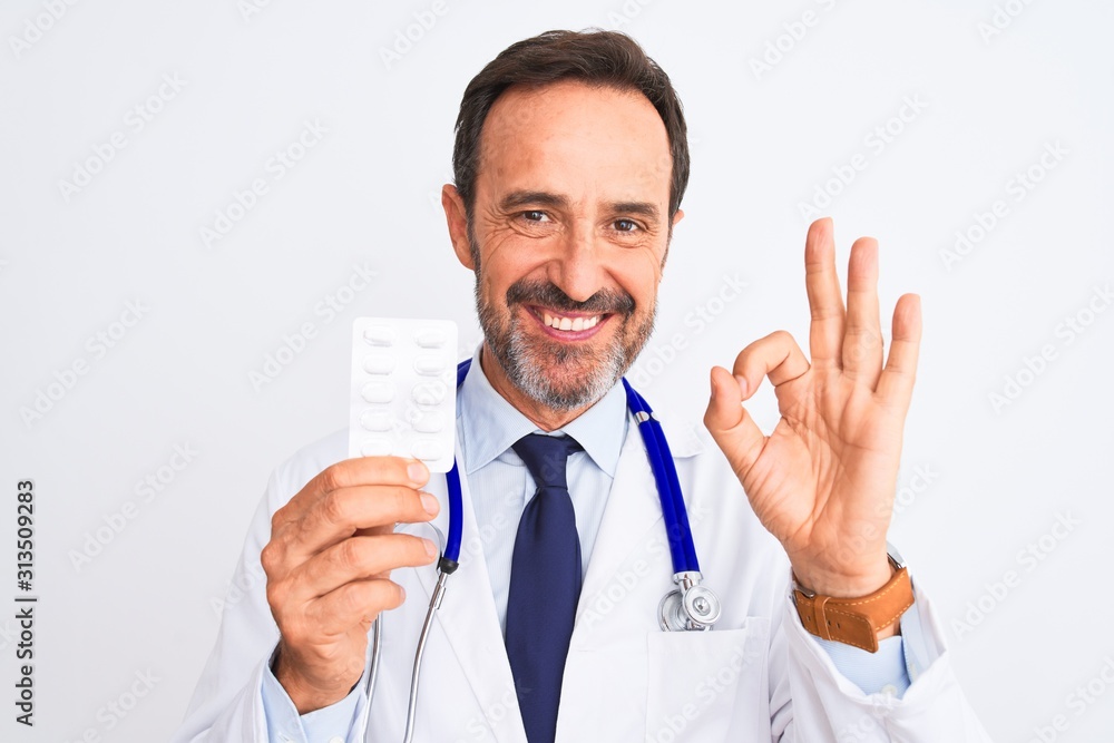 Middle age doctor man holding pills standing over isolated white background doing ok sign with fingers, excellent symbol