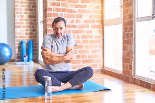 Middle age handsome sportman sitting on mat doing stretching yoga exercise at gym shaking and freezing for winter cold with sad and shock expression on face photo