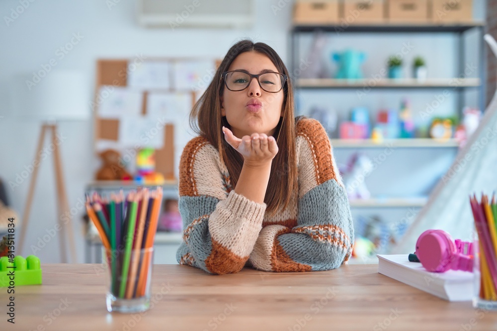 Young beautiful teacher woman wearing sweater and glasses sitting on desk at kindergarten looking at the camera blowing a kiss with hand on air being lovely and sexy. Love expression. Young beautiful teacher woman wearing sweater and glasses sitting on desk at kindergarten looking at the camera blowing a kiss with hand on air being lovely and sexy. Love expression.