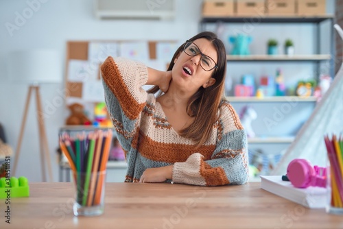 Young beautiful teacher woman wearing sweater and glasses sitting on desk at kindergarten Suffering of neck ache injury, touching neck with hand, muscular pain