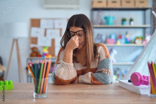 Young beautiful teacher woman wearing sweater and glasses sitting on desk at kindergarten tired rubbing nose and eyes feeling fatigue and headache. Stress and frustration concept.