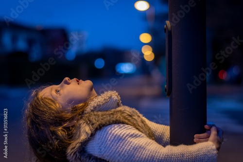 cute little girl is embracing a streetlight pylon and she is looking up to its light