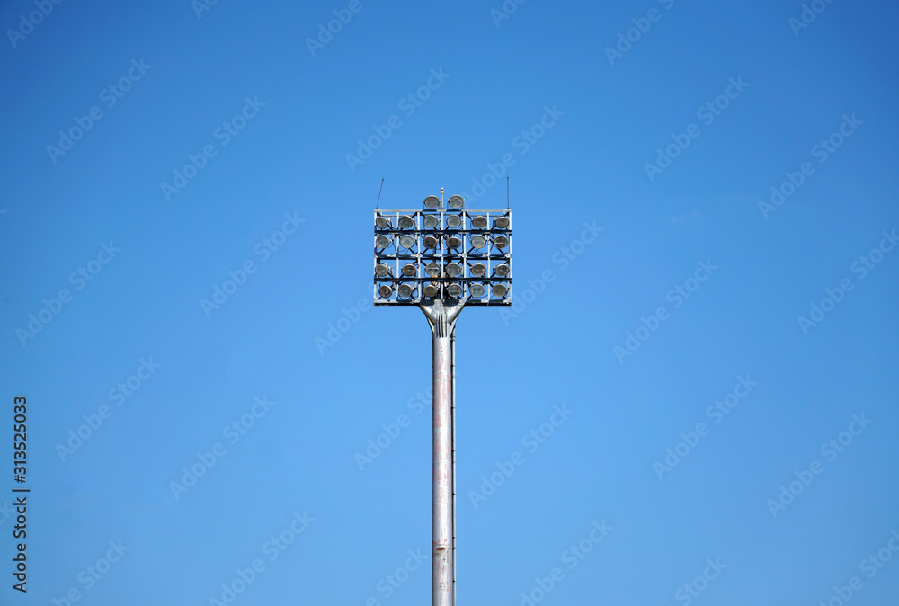 Spotlights tower with a metal pole for the sports arena. Installed around football stadium. Blue sky background. Spotlights tower with a metal pole for the sports arena. Installed around football stadium. Blue sky background.