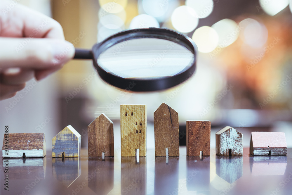 Hand holding magnifying glass and looking at house model, house selection, real estate concept. - obrazy, fototapety, plakaty 