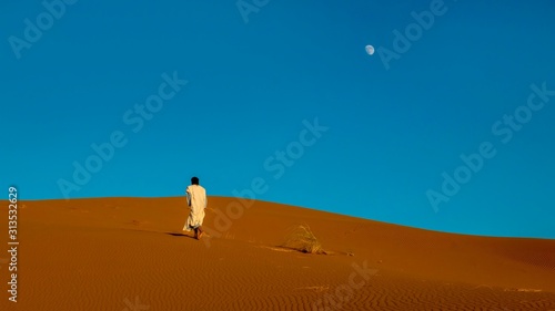 A Moroccan man wearing full length traditional nomadic clothing, walks barefoot uphill toward the ridge of a red sand dune in the Sahara Desert.