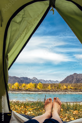 View from inside the tent on bare women's feet with copy space. Female hiker lying in tent with a view of mountain and river. Concept of travel and hiking.