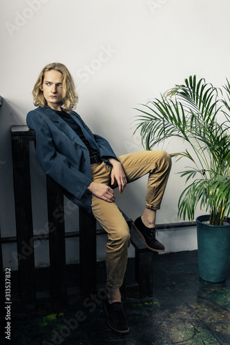 Young handsome guy with long blonde hair in a blue jacket poses, sits  on a dark background.