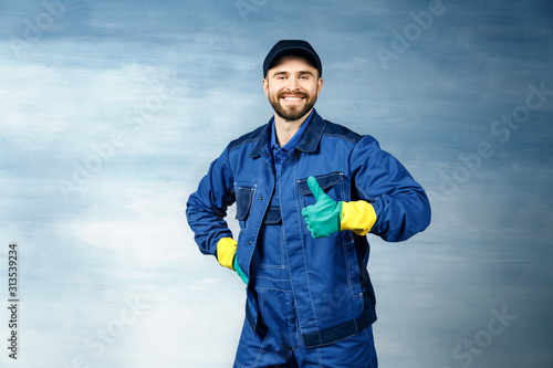 Thumb up. Young handsome man with a beard in a blue working uniform for cleaning rooms smiles isolated on blue background,thumbs up © Eno1