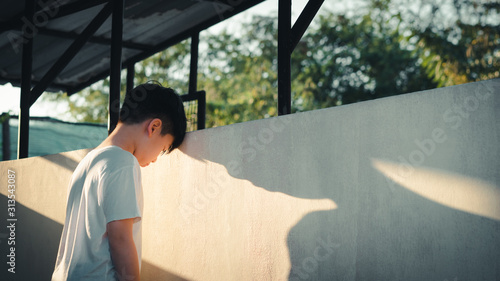 Depressed, frustrated and overwhelmed Asian teenage boy stand alone and lean his head against wall with casting shadow, feeling worthless, stressed, guilty, hopeless and having suicidal thought. photo