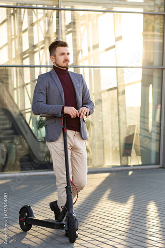 Young handsome stylish man with e-scooter standing on sidewalk near the business center on the sunset. Trendy urban transportation on modern electric scooter. Eco friendly mobility concept.