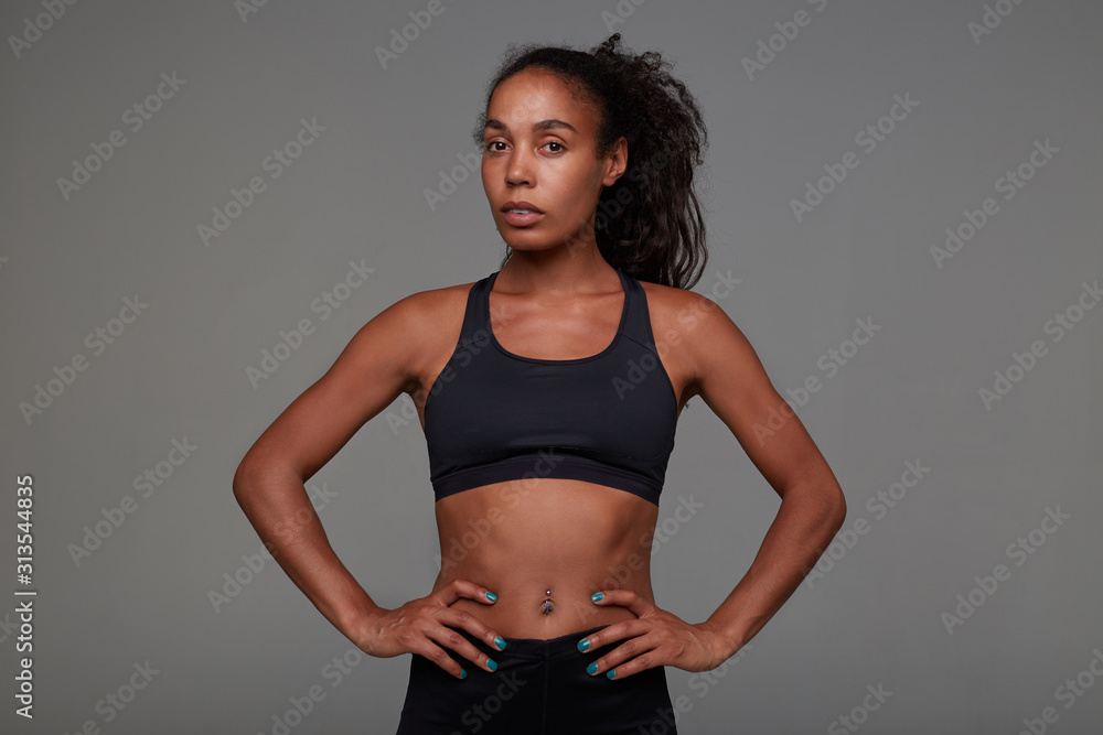 Studio shot of young pretty dark skinned curly lady with belly button piercing holding hands on her waist while posing over grey background in athletic black clothes Studio shot of young pretty dark skinned curly lady with belly button piercing holding hands on her waist while posing over grey background in athletic black clothes
