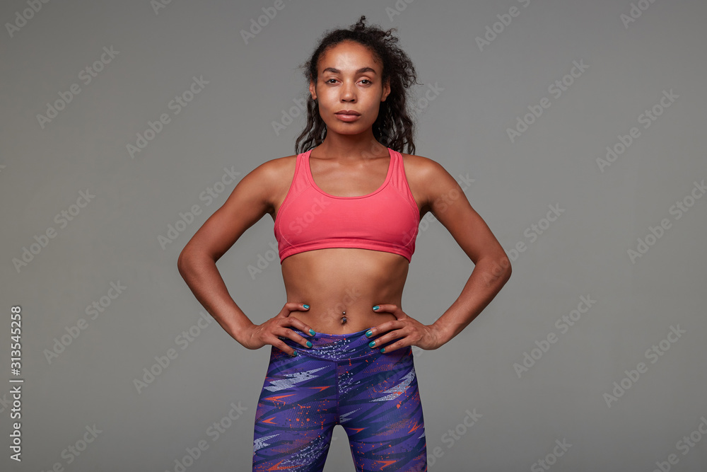 Serious young slim curly brunette woman in athletic wear holding hands on her waist while looking at camera with folded lips, standing against grey background Serious young slim curly brunette woman in athletic wear holding hands on her waist while looking at camera with folded lips, standing against grey background