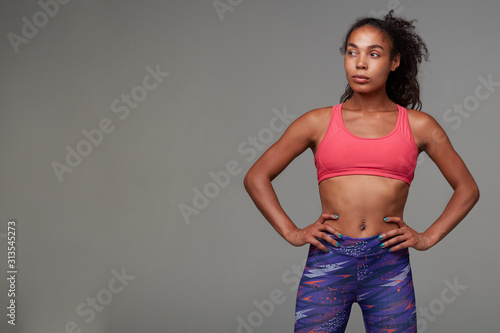 Studio photo of attractive young sporty dark skinned curly brunette lady keeping hands on her waist and looking seriously aside, isolated over grey background. Fitness male model