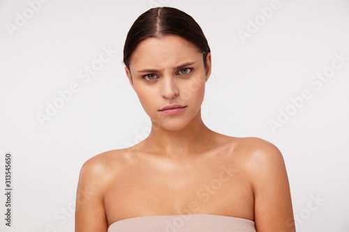 Portrait of indignant young dark haired green-eyed female with natural makeup looking severely at camera and frowning her eyebrows, standing against white background