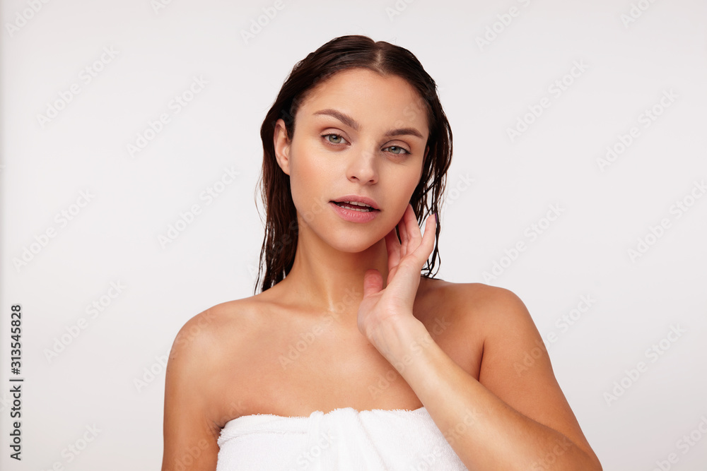 Indoor photo of charming young dark haired green-eyed lady without makeup touching soft her cheek and looking calmly at camera, posing over white background after having shower Indoor photo of charming young dark haired green-eyed lady without makeup touching soft her cheek and looking calmly at camera, posing over white background after having shower