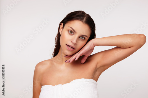 Attractive young brunette green-eyed female with wet hair touching gently her face with raised hand and looking affectionately to camera, posing over white background in bath towel